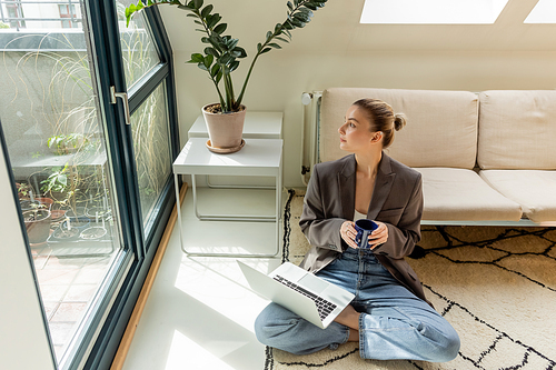 Young freelancer holding cup and looking at windows near laptop on carpet at home