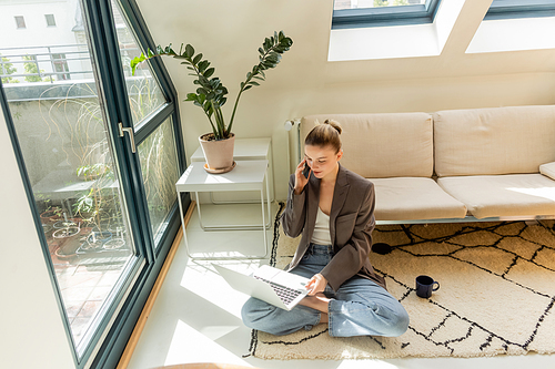 Young freelancer in jacket talking on smartphone and using laptop near cup on carpet at home