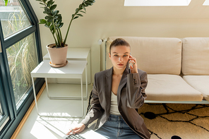 Young woman in blazer talking on smartphone and holding laptop in living room