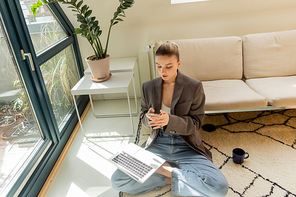 Young woman in jacket and jeans using smartphone and laptop near cup on carpet at home