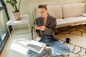Smiling freelancer using laptop and smartphone near cup on carpet on carpet