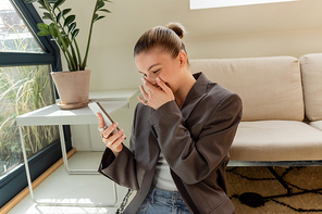 Cheerful woman in jacket using smartphone in living room