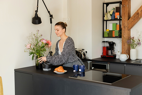 Woman in knitted cardigan putting vase with flowers near croissant and tea in kitchen