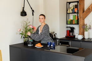 Smiling woman putting flowers in vase near cups and croissant in kitchen