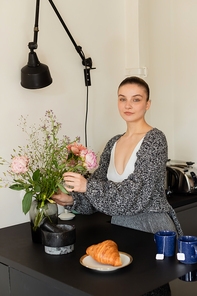 Woman in knitted cardigan putting flowers in vase near croissant and tea in kitchen