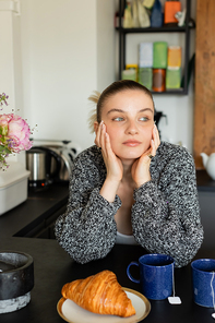 Woman in warm cardigan looking away near cups and blurred croissant in kitchen