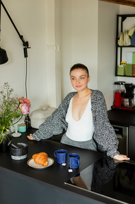 Woman in knitted cardigan standing near croissant and cups in kitchen