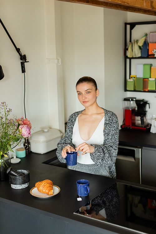 Young woman holding cup near croissant and flowers in kitchen