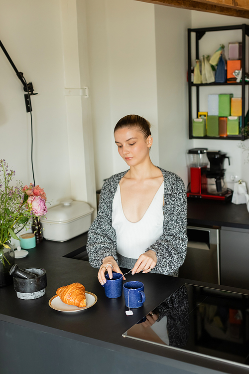 Woman in cardigan holding cup with tea bag near croissant in kitchen