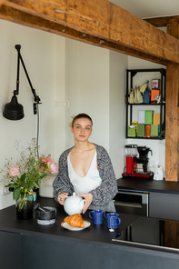 Young woman in knitted cardigan holding teapot near cups and croissant in kitchen