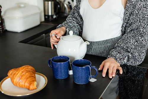 Cropped view of woman in knitted cardigan holding teapot near cups and blurred croissant at home