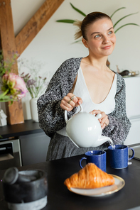 Positive woman in knitted cardigan pouring tea near croissant in kitchen