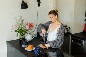 Woman in knitted cardigan cutting avocado near croissant and cups in kitchen