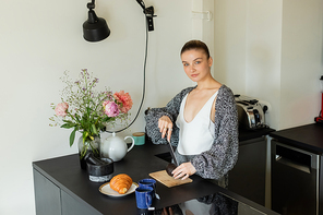 Young woman in warm cardigan cutting avocado near tea and croissant in kitchen