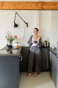 Young woman in knitted cardigan holding cup in modern kitchen at home
