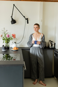 Positive woman holding cup and looking away near croissant in modern kitchen
