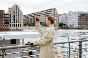 Side view of woman in trench coat taking photo on cellphone near river in Berlin