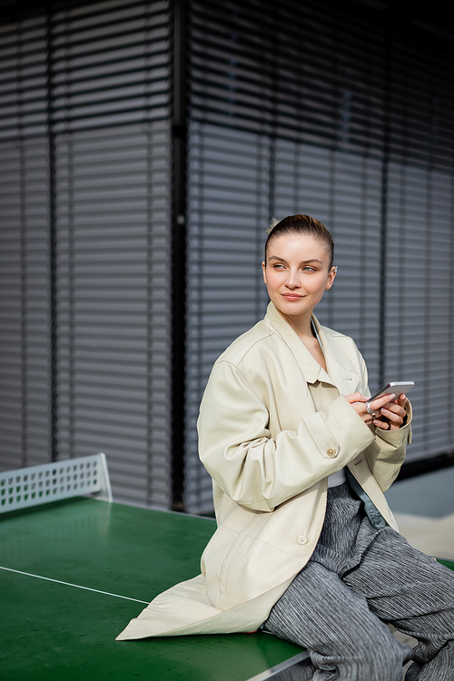 Woman in trench coat using smartphone while sitting on ping-pong table on street
