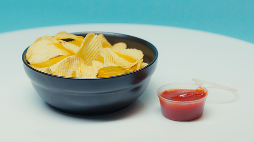 crunchy and ridged potato chips in bowl near plastic container with ketchup on blue