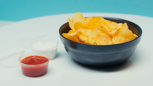 crunchy and ridged potato chips in bowl near plastic containers with ketchup and mayonnaise on blue