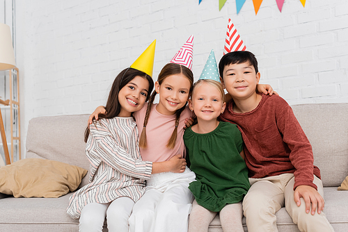Smiling interracial kids in party caps looking at camera and hugging during birthday party at home