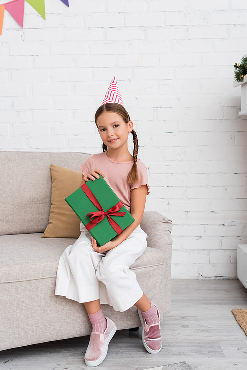 Cheerful girl in party cap holding gift box on couch at home