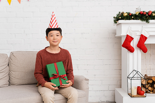Preteen asian boy in party cap holding present on couch near christmas decor at home