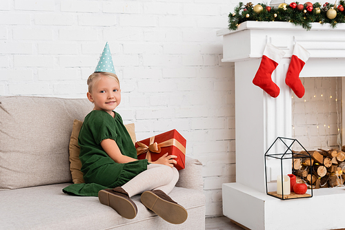 Smiling kid in party cap holding present during birthday party near christmas decor at home