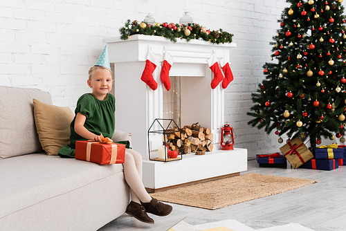 Preschooler girl in party cap holding gift near christmas decor and tree at home