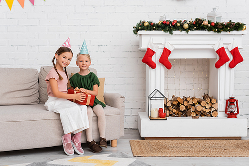 Smiling friends in party caps holding present on couch near christmas decor at home