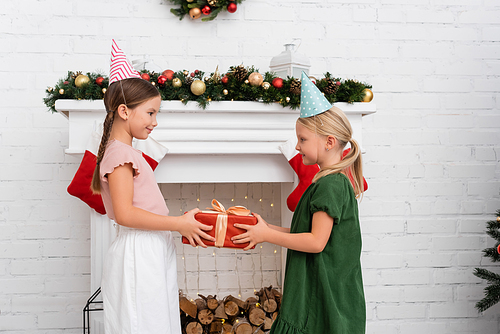 Side view of girl in party cap giving present to friend near christmas decor on fireplace at home