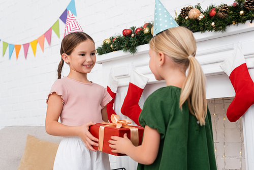 Smiling girl in party cap taking present from blurred friend near christmas decor at home