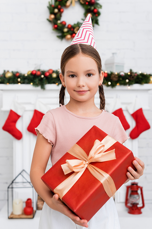 Portrait of kid in party cap holding gift box near blurred christmas decor at home