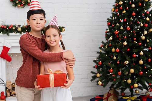 Asian boy in party cap hugging smiling friend with present near christmas tree at home