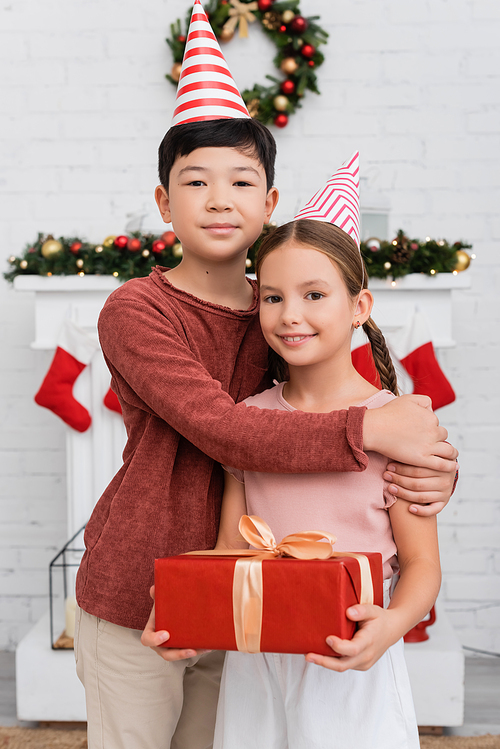 Asian boy hugging friend in party cap holding gift box near blurred christmas decor at home