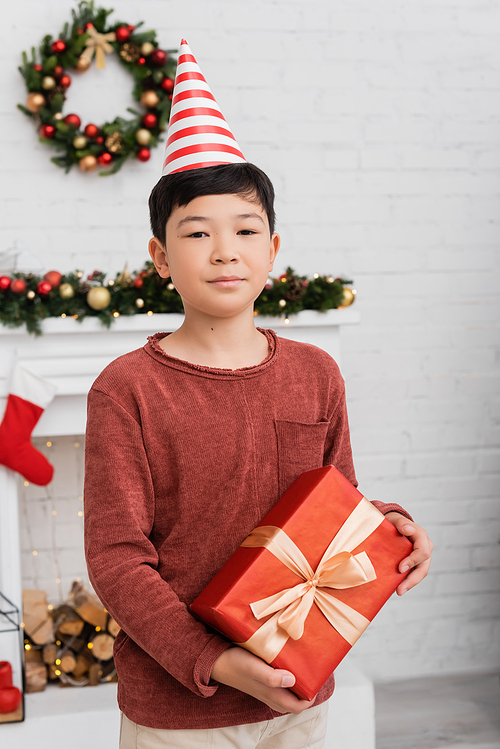Asian boy in party cap holding present and looking at camera near blurred christmas decor at home