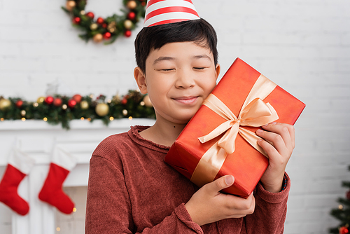 Happy asian boy in party cap holding gift near blurred christmas decor at home