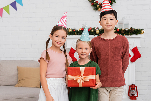 Multiethnic kids in party caps standing near friend with gift box and christmas decoration at home