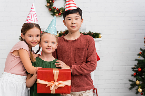 Interracial children in party caps hugging friend with present during birthday party at home