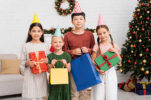 Multiethnic kids in party caps holding shopping bags and gifts near blurred christmas decor at home