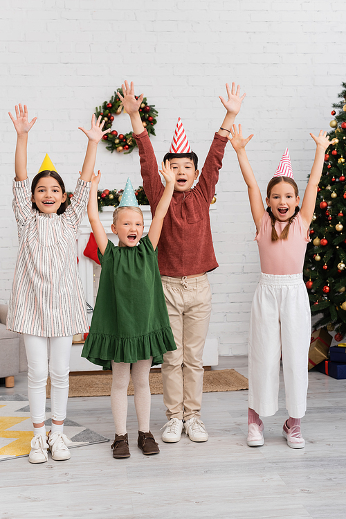 Cheerful interracial kids in party caps celebrating birthday party near blurred christmas decor at home