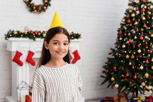 Smiling girl in party cap looking at camera near burred christmas tree at home