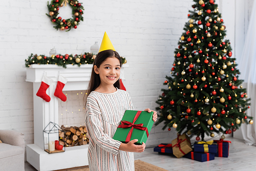 Happy girl in party cap holding gift box near blurred christmas tree and fireplace at home