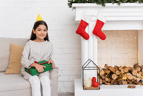 Smiling girl in party cap holding present on couch near fireplace with christmas decor at home