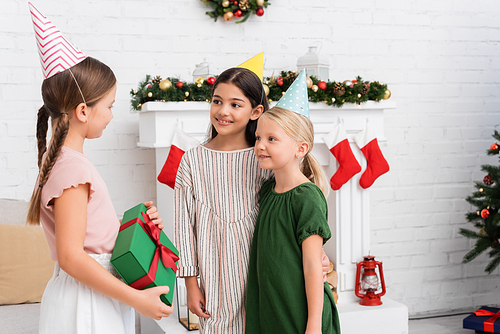 Positive girls in party caps looking at friend with gift near blurred christmas decor at home