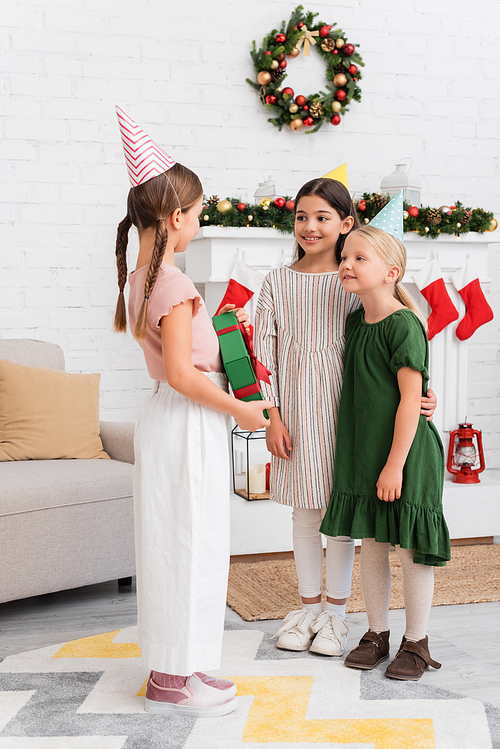 Girls in party caps looking at friend with gift box near blurred christmas stockings at home