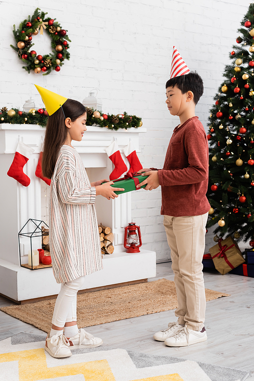 Multiethnic children in party caps holding present near christmas decor on fireplace at home