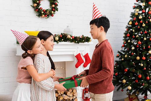 Asian boy in party cap giving present to cheerful friend near christmas decor on fireplace at home