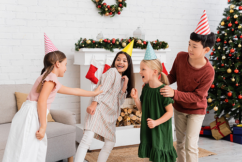 Cheerful interracial kids in party caps playing tag during birthday party near christmas decor at home