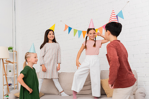 Cheerful girls in party caps looking at asian friend during birthday party at home